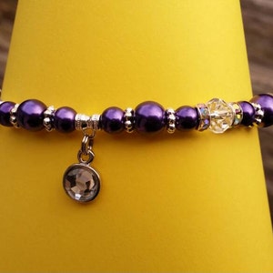 K&ouml;nnte beinhalten: Ein violettes und silbernes Perlenarmband mit einem Kristall-Anh&auml;nger. Das Armband liegt auf einem gelben Hintergrund.
