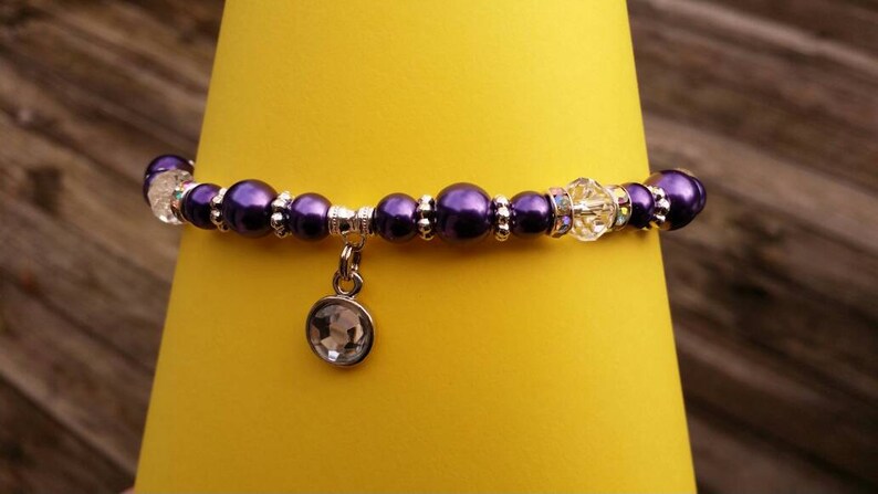 K&ouml;nnte beinhalten: Ein violettes und silbernes Perlenarmband mit einem Kristall-Anh&auml;nger. Das Armband liegt auf einem gelben Hintergrund.