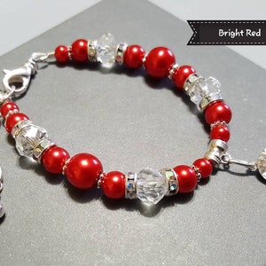 K&ouml;nnte beinhalten: Ein rotes und silbernes Armband mit einem Kristall-Anh&auml;nger. Das Armband besteht aus roten und silbernen Perlen und hat eine silberne Kette mit einem Karabinerverschluss. Der Anh&auml;nger ist ein gro&szlig;er Kristall mit einer silbernen Fassung.