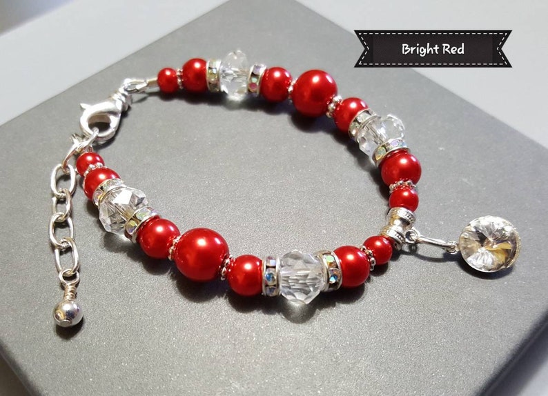 K&ouml;nnte beinhalten: Ein rotes und silbernes Armband mit einem Kristall-Anh&auml;nger. Das Armband besteht aus roten und silbernen Perlen und hat eine silberne Kette mit einem Karabinerverschluss. Der Anh&auml;nger ist ein gro&szlig;er Kristall mit einer silbernen Fassung.