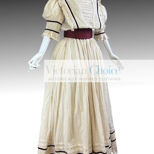 Pode incluir: Um vestido branco de algod&atilde;o com decote alto e mangas bufantes. O vestido tem uma faixa bord&ocirc; e detalhes pretos. O vestido &eacute; de estilo vitoriano e inspirado historicamente.