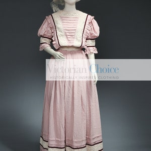 Pu&ograve; includere: Un abito in cotone rosa con un bordo in pizzo bianco e un accento di nastro bordeaux. L'abito ha un corpetto aderente e una gonna ampia. L'abito &egrave; indossato da un manichino con un cappello bianco.