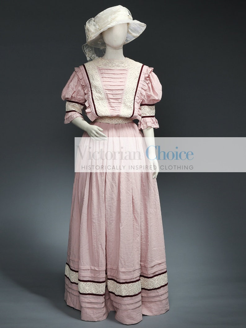 Pu&ograve; includere: Un abito in cotone rosa con un bordo in pizzo bianco e un accento di nastro bordeaux. L'abito ha un corpetto aderente e una gonna ampia. L'abito &egrave; indossato da un manichino con un cappello bianco.