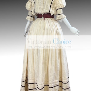 Pode incluir: Um vestido de algod&atilde;o branco com uma faixa bord&ocirc; e detalhes em renda. O vestido tem um corpete justo e uma saia volumosa. O vestido &eacute; de estilo vitoriano.