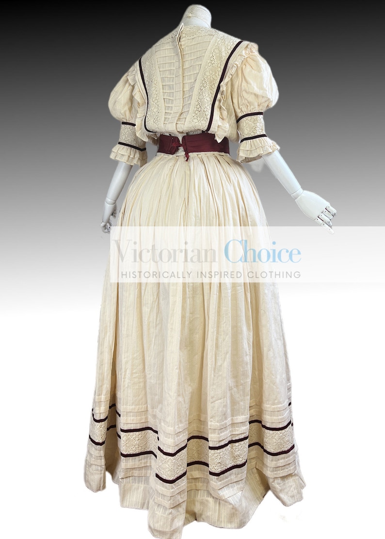 Pode incluir: Um vestido de algod&atilde;o branco com uma faixa bord&ocirc; e detalhes em renda. O vestido tem um corpete justo e uma saia volumosa. O vestido &eacute; de estilo vitoriano.