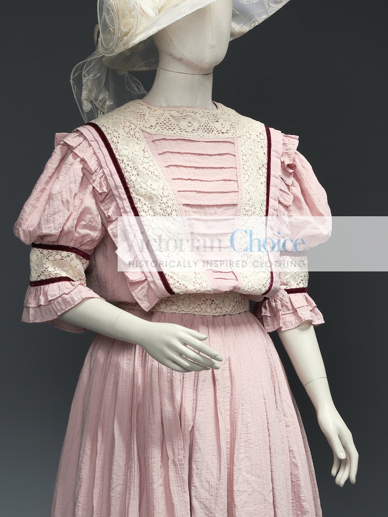 Pu&ograve; includere: Un abito in cotone rosa con un bordo in pizzo e un nastro bordeaux. L'abito ha un corpetto aderente e una gonna ampia. L'abito &egrave; indossato da un manichino con un cappello bianco.