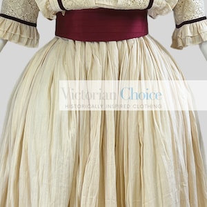 Pode incluir: Um vestido cor de creme com uma faixa bord&ocirc; e detalhes em renda. O vestido tem uma saia cheia e &eacute; feito de um tecido leve. Victorian Choice, Roupas de inspira&ccedil;&atilde;o hist&oacute;rica.