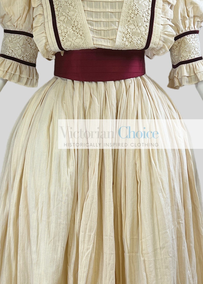 Pode incluir: Um vestido cor de creme com uma faixa bord&ocirc; e detalhes em renda. O vestido tem uma saia cheia e &eacute; feito de um tecido leve. Victorian Choice, Roupas de inspira&ccedil;&atilde;o hist&oacute;rica.