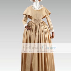May include: A beige Victorian era dress with a white lace collar and a matching cape. The dress has a fitted bodice and a full skirt. The model is wearing a white lace bonnet.