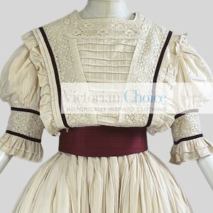 Pode incluir: Um vestido branco de estilo vitoriano com um corpete de renda e uma faixa bord&ocirc;. O vestido tem uma saia cheia e mangas bufantes com detalhes em renda.