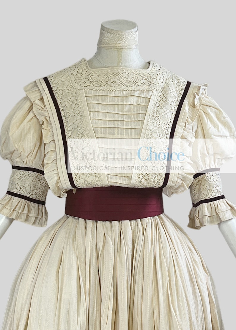 Pode incluir: Um vestido branco de estilo vitoriano com um corpete de renda e uma faixa bord&ocirc;. O vestido tem uma saia cheia e mangas bufantes com detalhes em renda.