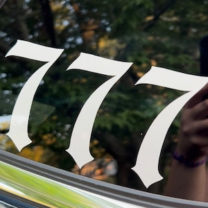 以下が含まれることがあります： スタイリッシュなフォントで777という数字が書かれた白いビニールステッカー。ステッカーは車の窓に貼られています。