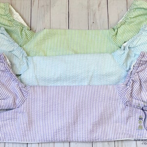 May include: Three pairs of baby bloomers in different colors: light green, light blue, and lavender. Each pair has a white stripe pattern and a ruffled edge.