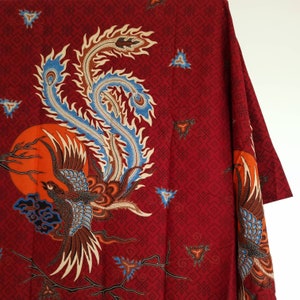 Può includere: Tessuto rosso con un motivo batik tradizionale che presenta una fenice con accenti blu e arancioni. Lo sfondo è un motivo geometrico ripetitivo.