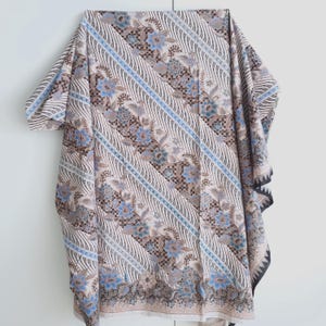 Puede incluir: Una tela estampada en beige, azul y marrón con un diseño floral. La tela está drapeada, mostrando un patrón repetitivo de flores y rayas diagonales. Los bordes tienen un borde decorativo. La tela parece ser un chal o una prenda similar.