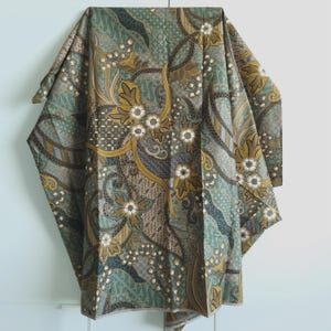 Puede incluir: Textil doblado con un detallado estampado floral en tonos verdes, marrones y dorados. El diseño presenta flores estilizadas y espirales, creando una apariencia rica y texturizada. La tela está drapeada, mostrando el intrincado patrón.