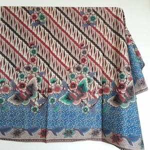 Puede incluir: Una tela estampada con un diseño diagonal repetido en crema, rojo y marrón. El borde inferior tiene un borde azul con motivos florales y de mariposas en tonos verdes, rojos y grises. La tela está doblada.