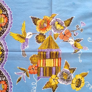Peut inclure: Textile bleu clair avec un motif floral et aviaire vibrant. Le motif comprend des fleurs jaunes, orange, violettes et marron, ainsi que des colibris et une maison stylisée. La bordure présente un motif décoratif violet et noir.
