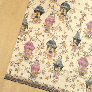 Könnte beinhalten: Weißer Stoff mit einem sich wiederholenden Muster aus violetten und blauen Vogelkäfigen mit floralen Akzenten. Die Vogelkäfige sind mit kleinen Vögeln und Blumen gefüllt. Der Hintergrund ist cremefarben.