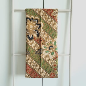 以下が含まれることがあります： 長方形のバティック生地。花柄が特徴で、金、黒、白の色合いの大きな花が、緑、赤、茶色の背景に描かれています。白い梯子に生地が掛けられています。