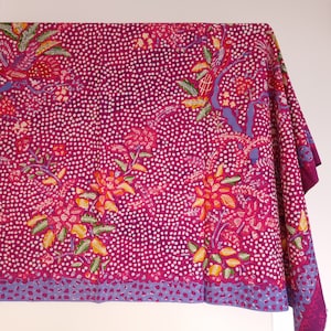 Op de afbeelding: Een magenta stof met witte stippen en bloemmotieven. De bloemen zijn rood, oranje en geel, met groene en blauwe accenten. Een blauwe rand loopt langs de onderkant. Geschikt voor tafelkleden of decoraties.