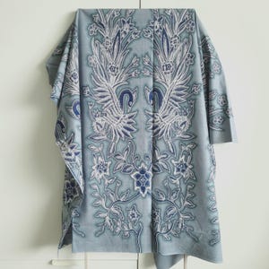 Può includere: Un tessuto azzurro con un motivo simmetrico di fiori e uccelli bianchi e blu. Il tessuto è drappeggiato, rivelando i dettagli della stampa. Il tema è il design tessile, con motivi intricati.