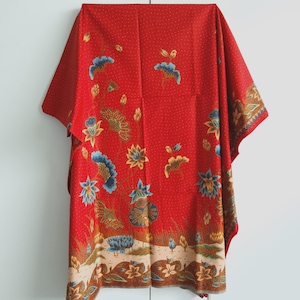 Può includere: Un tessuto rosso con un motivo floreale. Il design presenta fiori e foglie blu, gialli e marroni. Il tessuto ha uno sfondo a pois e un bordo beige con altri motivi floreali. L'indumento sembra essere un top o un vestito ampio.