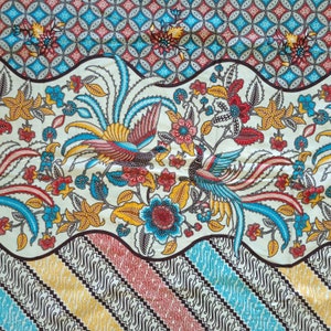 Op de afbeelding: Een witte stof met een blauw, rood en geel bloemenpatroon. De stof heeft twee gestileerde vogels met lange staarten.