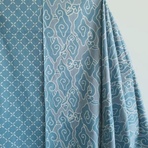 Peut inclure: Deux sections de tissu avec des motifs différents. La section de gauche a un fond bleu clair avec un motif géométrique blanc. La section de droite a un fond bleu clair avec un motif en forme de nuage blanc. Le tissu est drapé.