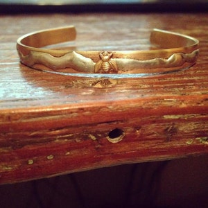 Könnte beinhalten: Ein gold- und silberfarbenes Armband mit einem Bienen-Design. Das Armband ist aus Metall und hat eine gehämmerte Textur.