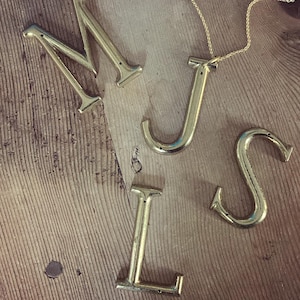 以下が含まれることがあります： 「MISS」という単語を綴る金色の金属文字と、文字「J」に金色のチェーンネックレスが取り付けられています。