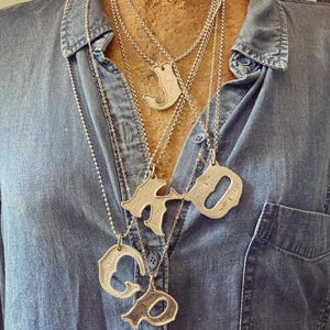 May include: Silver initial pendant necklaces displayed on a denim shirt. The necklaces feature capital letters with decorative detailing. The chains are delicate and the pendants are textured.