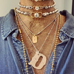 May include: A collection of layered necklaces in silver and gold tones. The necklaces include beaded chains, pendants with the letter "J" and a decorative "D", and chains with clear crystal accents. The necklaces are displayed on a wooden bust, against a denim shirt.