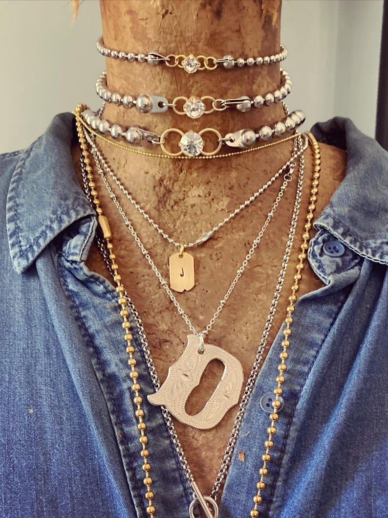 May include: A collection of layered necklaces in silver and gold tones. The necklaces include beaded chains, pendants with the letter "J" and a decorative "D", and chains with clear crystal accents. The necklaces are displayed on a wooden bust, against a denim shirt.