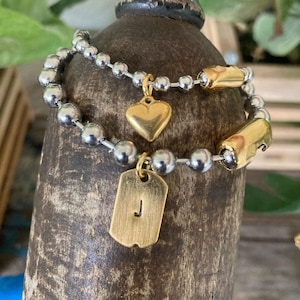 May include: A two-strand beaded bracelet featuring silver-toned ball chains, gold-toned cylindrical accents, a small gold-toned heart charm, and a rectangular gold-toned dog tag pendant engraved with the letter J.
