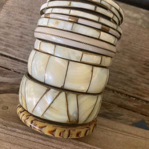 May include: A stack of assorted bangles featuring various designs, including cream-colored segments with metallic accents, smooth light-toned bands, and a single bottom bangle with a brown and cream marbled pattern, all arranged in a vertical column.