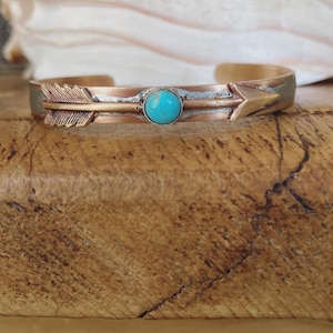 Puede incluir: Un brazalete de flecha de cobre y plata con una piedra turquesa en el centro. La flecha apunta hacia la derecha.