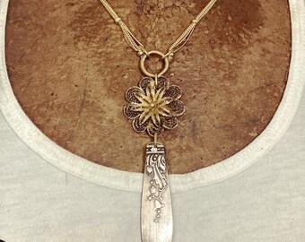May include: A gold-toned necklace with a filigree flower pendant and a silver spoon-shaped charm. The necklace has multiple strands and a circular clasp. The spoon charm has an ornate floral design. The word "VINTAGE" is visible on the shirt.