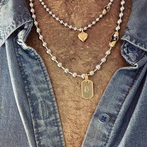 May include: A layered necklace set featuring two silver-toned ball chains. The shorter chain has a small gold-toned heart charm. The longer chain features a gold-toned rectangular dog tag pendant engraved with the letter B and gold-toned accents.