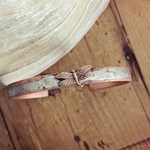 Könnte beinhalten: Ein Armband aus Kupfer und Silber mit einem Libellen-Anhänger. Die Libelle ist aus Messing und hat eine goldene Oberfläche.