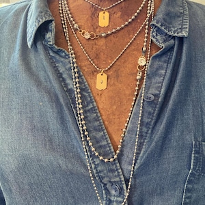 May include: A collection of layered necklaces with varying chain styles. The necklaces include ball chains, delicate chains, and chains with small, round beads. Some necklaces feature gold-colored dog tag pendants with the letter "J" and "B".