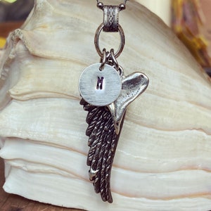 May include: A silver necklace with a silver charm in the shape of a wing. The charm has a small round disc attached to it with the letter "M" engraved on it.