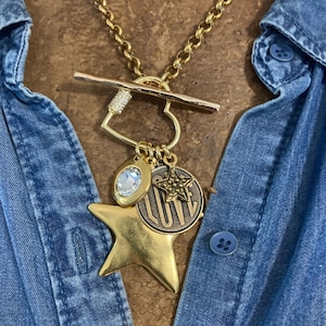 May include: Gold-toned necklace with a toggle clasp and multiple charms. Charms include a star, a coin with the word "LUV", a star-shaped charm, and a teardrop-shaped charm. The necklace is worn over a blue denim shirt.