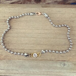 May include: An anklet featuring a silver ball chain with a gold-colored clasp. A small gold-colored charm with a clear crystal is centered on the chain. The anklet is displayed on a wooden surface.