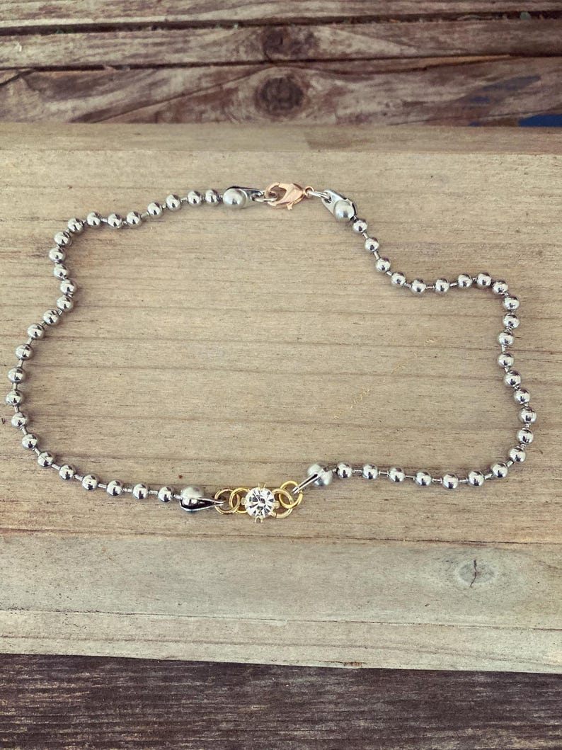 May include: An anklet featuring a silver ball chain with a gold-colored clasp. A small gold-colored charm with a clear crystal is centered on the chain. The anklet is displayed on a wooden surface.