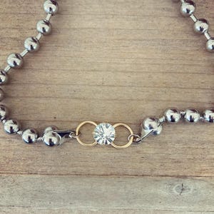 May include: Silver ball chain necklace with a gold clasp and a central gold accent holding a clear, sparkling gemstone. The necklace is displayed on a wooden surface, showcasing its reflective qualities and design.