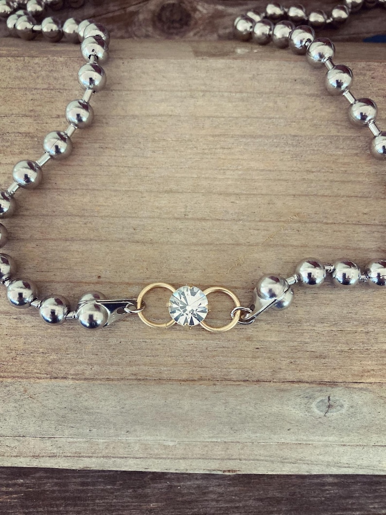 May include: Silver ball chain necklace with a gold clasp and a central gold accent holding a clear, sparkling gemstone. The necklace is displayed on a wooden surface, showcasing its reflective qualities and design.