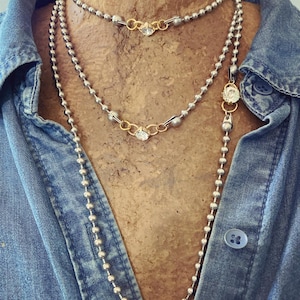 May include: Three silver necklaces with gold accents and clear gemstones. The necklaces are layered, with one choker-style, one mid-length, and one long chain. The necklaces are displayed on a mannequin wearing a blue denim shirt.