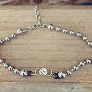 May include: A silver-toned necklace featuring a series of small, round beads. The necklace has a central design element with a clear, round gemstone set between two gold-toned rings. The necklace is adjustable with a chain clasp.