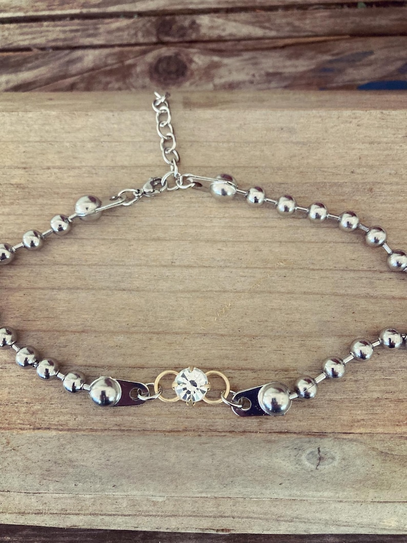 May include: A silver-toned necklace featuring a series of small, round beads. The necklace has a central design element with a clear, round gemstone set between two gold-toned rings. The necklace is adjustable with a chain clasp.
